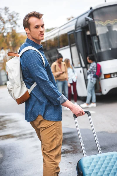 Selective focus of handsome man with backpack carrying wheeled bag near travel bus at street — Stock Photo