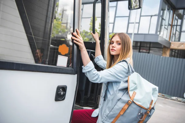 Attractive young female tourist with backpack walking into travel bus at urban street — Stock Photo