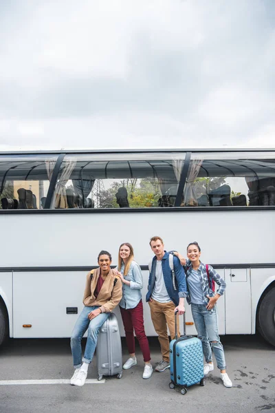 Giovani amici multiculturali con borse a ruote in posa vicino autobus di viaggio in strada — Foto stock