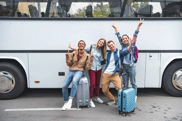 Turistas multiétnicos alegres con bolsas de viaje haciendo señales de paz cerca de autobús en la calle - foto de stock