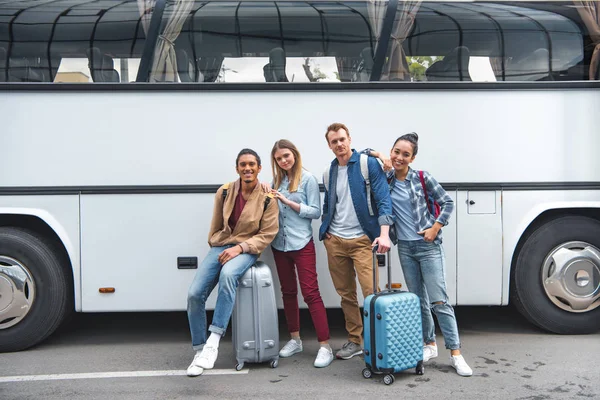 Junge multikulturelle Freunde mit Rolltaschen posieren in der Nähe des Reisebusses an der Stadtstraße — Stockfoto
