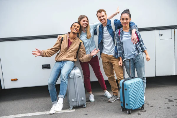 Amigos felices multiétnicos con bolsas de viaje gestos cerca de autobús en la calle - foto de stock