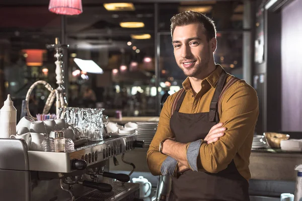 Beau jeune barista avec les bras croisés debout près de la machine à café et regardant la caméra — Photo de stock