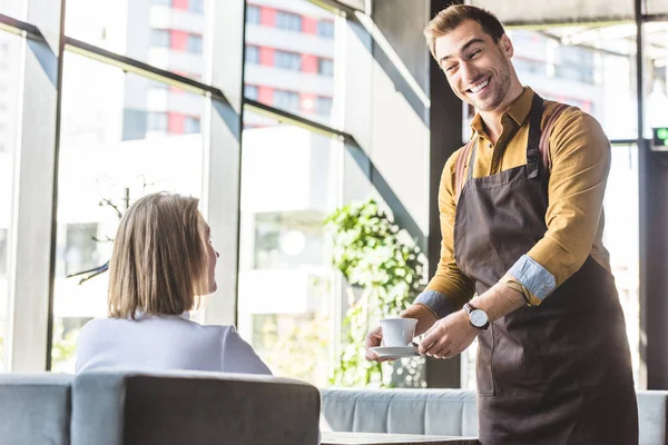 Wide smiling young waiter serving cup of coffee for female client at cafe — Stock Photo