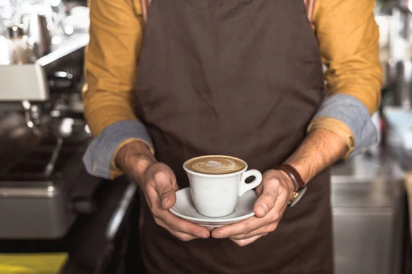 Cropped shot of barista in apron holding cup of fresh made cappuccino — Stock Photo