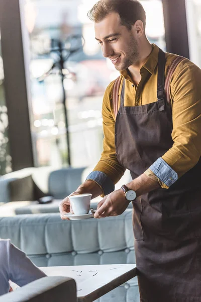 Sonriente joven camarero sirviendo taza de café para el cliente en la cafetería - foto de stock