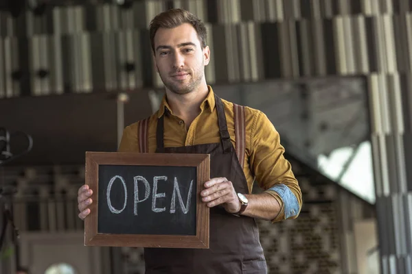 Attractive young waiter holding board with open inscription in cafe — Stock Photo