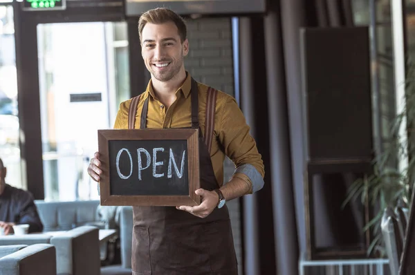 Handsome young waiter holding board with open inscription in cafe — Stock Photo