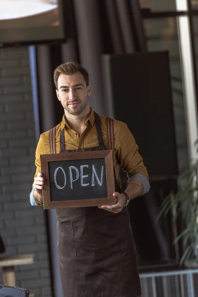 Handsome waiter in apron holding chalkboard with open inscription in cafe — Stock Photo