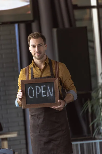 Beau garçon dans tablier tenant tableau avec inscription ouverte dans le café — Photo de stock