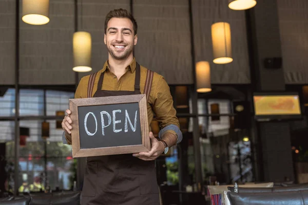 Attractive smiling waiter holding chalkboard with open inscription in cafe — Stock Photo