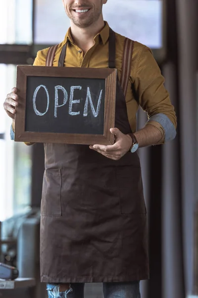 Plan recadré de jeune garçon souriant dans un tablier tenant un tableau noir avec inscription ouverte dans un café — Photo de stock