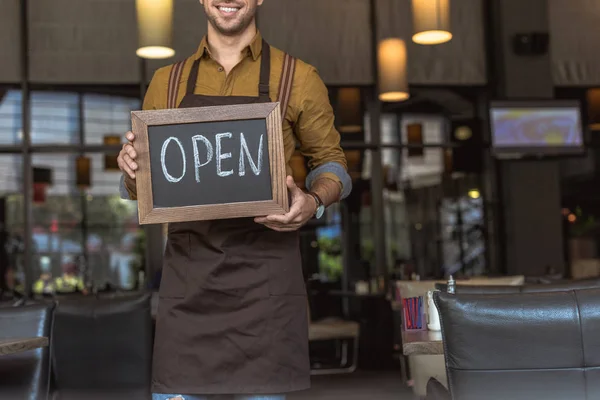 Tiro recortado de garçom feliz segurando quadro com inscrição aberta no café — Fotografia de Stock