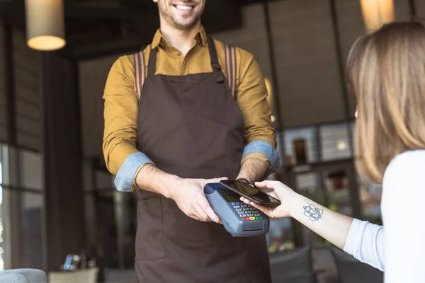 Plan recadré de serveur souriant tenant terminal de paiement tandis que le client faisant l'achat sans contact avec smartphone dans le café — Photo de stock