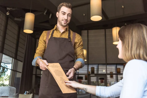 Bonito jovem garçom mostrando lista de menu para cliente feminino no café — Fotografia de Stock