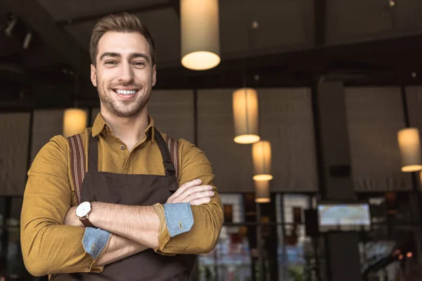 Усміхнений молодий офіціант з схрещеними руками дивиться на камеру в кафе — стокове фото
