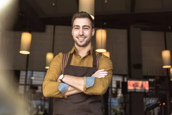 Schöner junger Kellner mit verschränkten Armen, der im Café in die Kamera schaut — Stockfoto