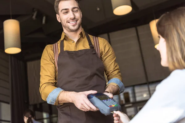 Feliz joven camarero sosteniendo terminal de pago mientras el cliente inserta la tarjeta de crédito - foto de stock