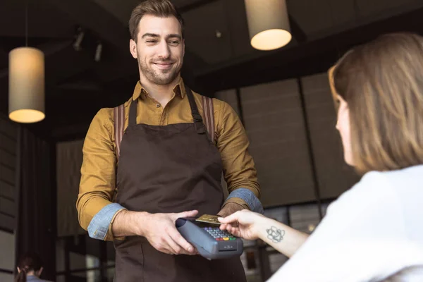 Счастливый молодой официант держит платежный терминал, пока клиент делает бесконтактную покупку с помощью кредитной карты в кафе — стоковое фото