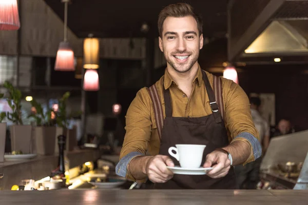 Sonriente joven camarero sosteniendo taza de café y mirando a la cámara - foto de stock