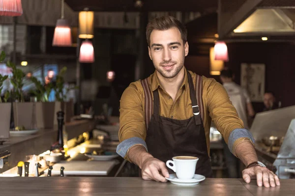 Atractivo joven barista con taza de delicioso café mirando a la cámara - foto de stock