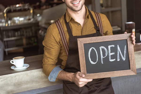 Cropped shot of happy young waiter holding chalkboard with open inscription in cafe — Stock Photo