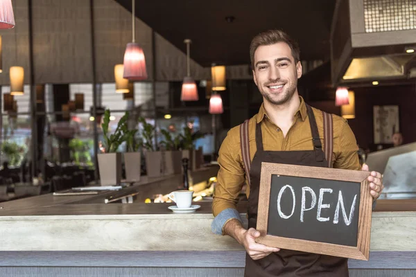 Feliz joven camarero sosteniendo pizarra con inscripción abierta en la cafetería - foto de stock