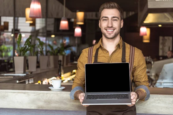 Feliz joven camarero sosteniendo portátil con pantalla en blanco en la cafetería - foto de stock