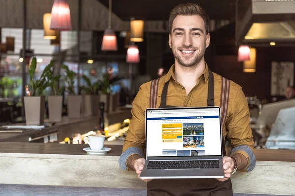 Apuesto joven camarero sosteniendo portátil con sitio web de reserva en la pantalla en la cafetería - foto de stock