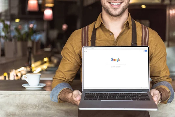 Cropped shot of smiling young waiter holding laptop with google website on screen in cafe — Stock Photo