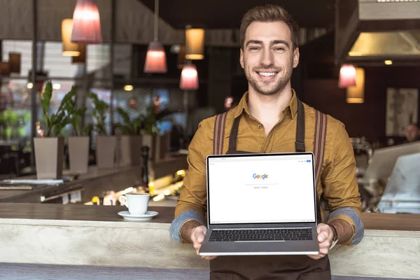 Apuesto joven camarero sosteniendo portátil con google sitio web en la pantalla en la cafetería - foto de stock