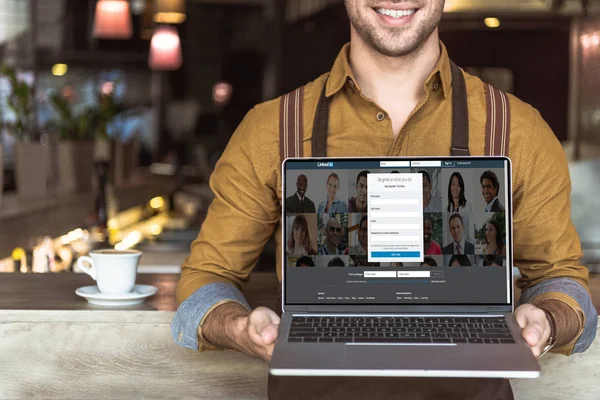 Обрезанный снимок улыбающегося молодого официанта, держащего ноутбук со ссылкой на сайт на экране в кафе — стоковое фото
