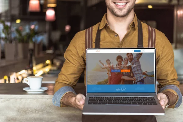 Cropped shot of smiling young waiter holding laptop with couchsurfing website on screen in cafe — Stock Photo