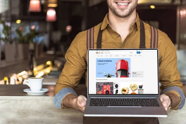 Tiro recortado de garçom jovem sorridente segurando laptop com site ebay na tela no café — Fotografia de Stock