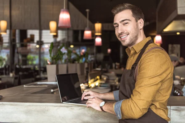 Beau jeune serveur en utilisant un ordinateur portable un comptoir de bar dans le café — Photo de stock