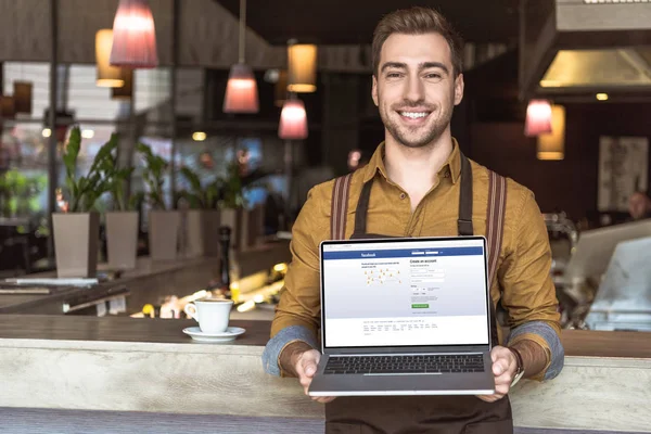 Apuesto joven camarero sosteniendo portátil con facebook sitio web en la pantalla en la cafetería - foto de stock