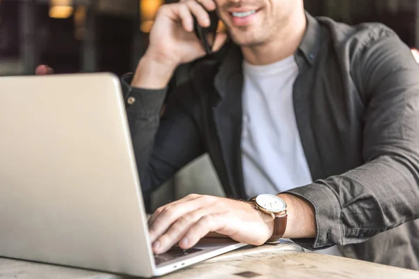 Обрізаний знімок усміхненого молодого фрілансера, який працює з ноутбуком і розмовляє по телефону в кафе — стокове фото