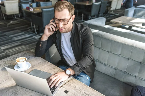 Confiant jeune pigiste travailler avec ordinateur portable et parler par téléphone dans le café — Photo de stock