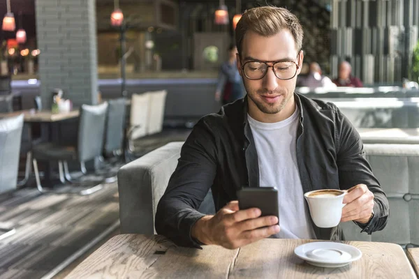Atractivo joven con taza de café usando teléfono inteligente en la cafetería - foto de stock