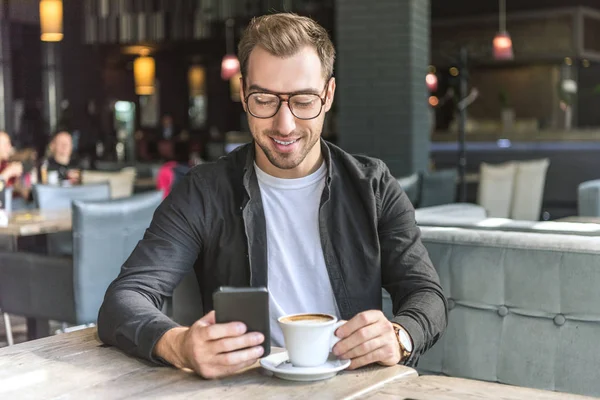 Jovem sorridente com xícara de café usando smartphone no café — Fotografia de Stock