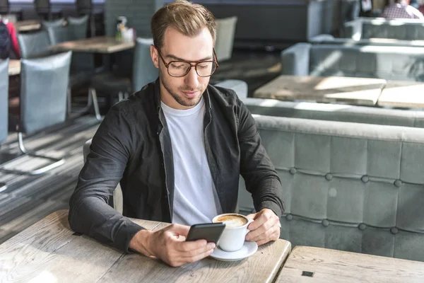 Beau jeune homme avec une tasse de café en utilisant un smartphone dans le café — Photo de stock