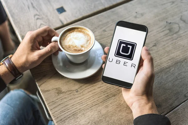 Recortado disparo de hombre con taza de cappuccino usando teléfono inteligente con el logotipo uber en la pantalla - foto de stock