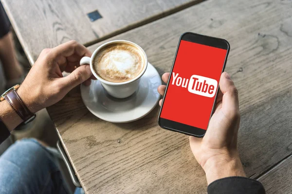 Recortado disparo de hombre con taza de cappuccino usando teléfono inteligente con logotipo de youtube en la pantalla — Stock Photo