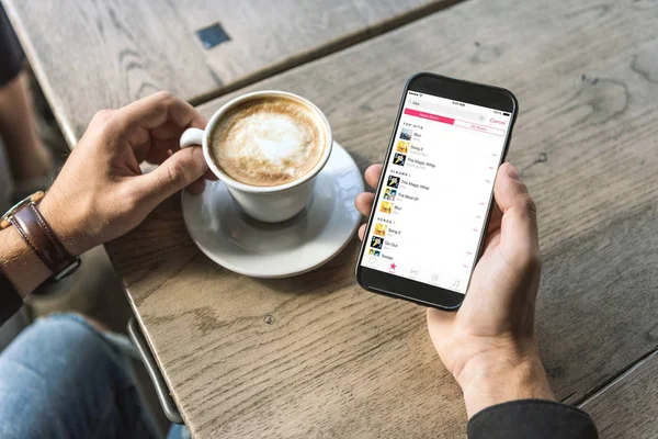 Colpo ritagliato di uomo con tazza di cappuccino utilizzando smartphone con musica mela sullo schermo — Foto stock