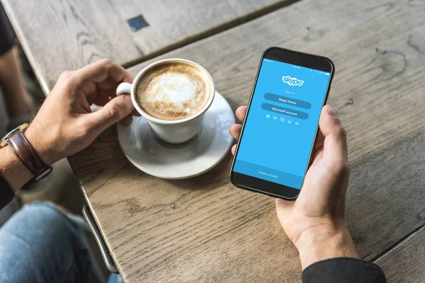 Recortado disparo de hombre con taza de cappuccino utilizando el teléfono inteligente con aplicación de Skype en la pantalla - foto de stock