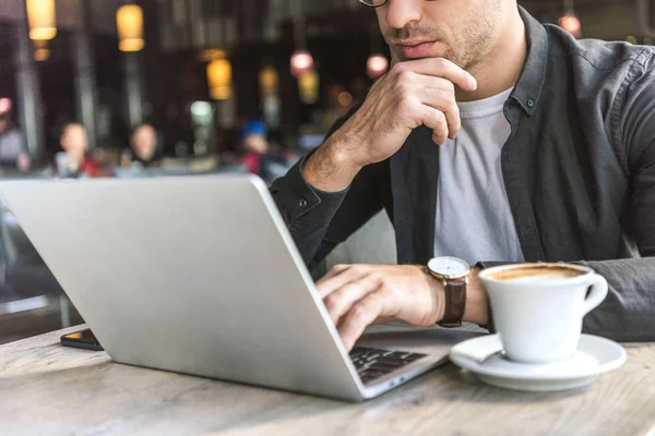 Recortado disparo de joven freelancer trabajando con el ordenador portátil en la cafetería con taza de café en la mesa - foto de stock