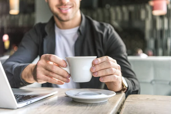 Plan recadré de jeune pigiste avec ordinateur portable tenant tasse de café dans le café — Photo de stock