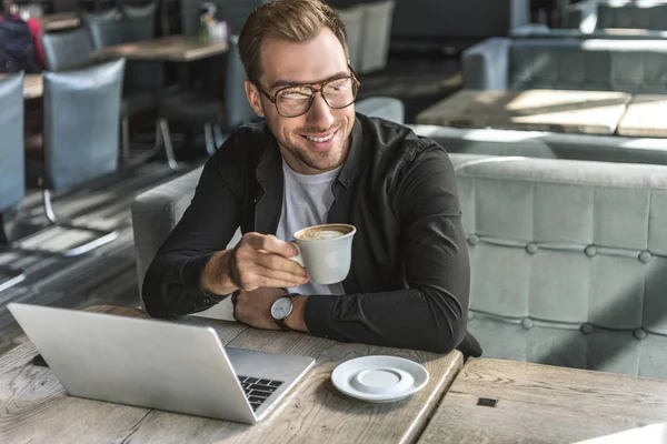 Bel giovane freelance con tazza di caffè e laptop seduto nel caffè — Foto stock