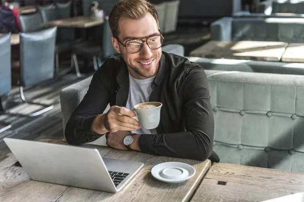 Bonito jovem freelancer com xícara de café e laptop sentado no café — Fotografia de Stock