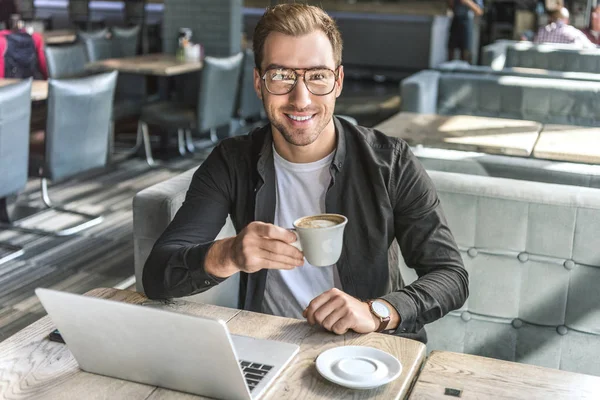 Jovem freelancer bonito com xícara de café e laptop olhando para a câmera no café — Fotografia de Stock